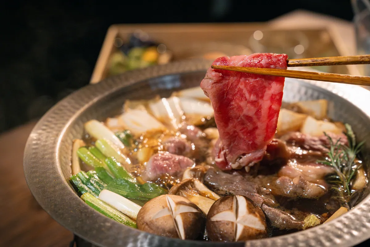 箸で持ち上げた牛肉と、ねぎ、しいたけが入った京風すき焼きの鍋