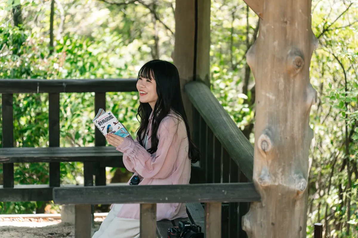 木のベンチに座り、野鳥図鑑を手に森を眺める女性