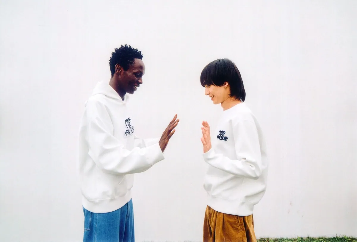 胸に刺繍ロゴの入った白いフーディーとプルオーバーを着た二人が、手を合わせて笑い合う様子