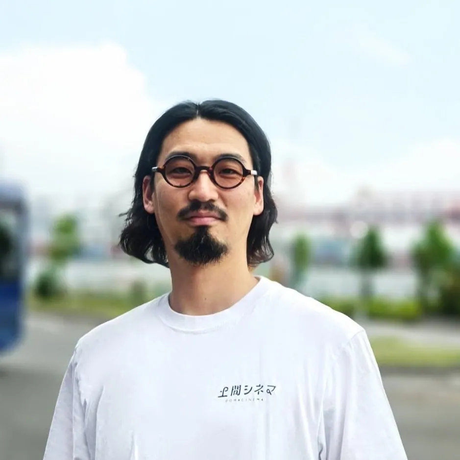 丸いメガネをかけ、土間シネマのロゴ入りTシャツを着た吉田直史館長