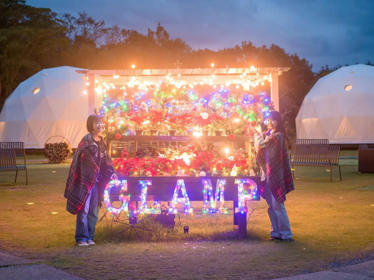 Two people wrapped in blankets standing in front of a welcome sign lit with Christmas lights, with dome tents behind them